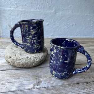 May include: Two ceramic mugs with a dark blue speckled glaze. The mugs have a rounded shape with a handle and a slightly flared rim. One mug rests on a gray stone, while the other sits on a wooden surface. The mugs are similar in size and design.