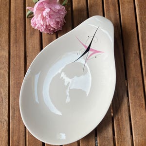 May include: Cream-colored, mid-century modern serving dish with a teardrop shape. The dish features a black and pink starburst design. A pink peony flower rests on a wooden surface next to the dish.