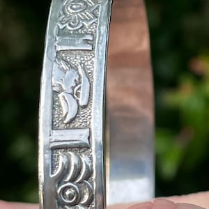 May include: Silver bangle bracelet with embossed decorative patterns. The bracelet features repeating geometric and floral designs, along with stylized figures. The details are raised against a textured background. The bracelet is shown against a blurred green background.