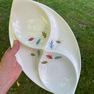 May include: A white, three-section serving dish with a leaf design in red, blue, and green. The dish has a unique, elongated shape and is held against a green background.