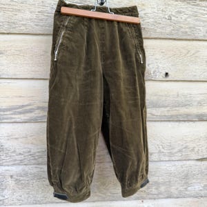 May include: Olive green corduroy pants with a high waist and a gathered, elasticized hem. The pants have two zippered pockets and are hanging on a wooden hanger against a weathered wood background.