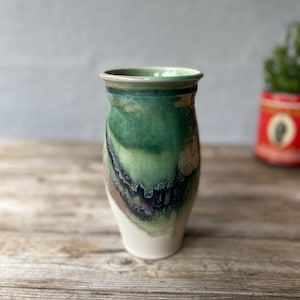 May include: A handmade ceramic vase with a unique, organic design. The vase features a blend of green, brown, and white colors, with a textured, layered appearance. The vase is approximately 7 inches tall and has a wide opening.