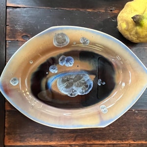 May include: A large, oval-shaped ceramic serving dish with a light brown glaze. The dish has a dark blue and brown swirl design with white speckles.