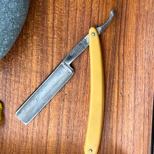 May include: A vintage straight razor with a yellow handle and a silver blade. The blade is open, revealing the sharp edge. The text "Otto Deutsch's HANS" is visible on the blade. The razor is set against a wooden surface.