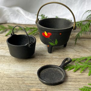 May include: Three miniature cast iron cookware pieces: a pot with a brass handle and a painted red flower, a smaller pot with a handle, and a small frying pan. The cookware is on a wooden surface.