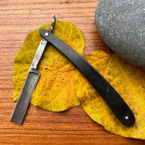 May include: A vintage straight razor with a black handle and silver blade, resting on a yellow leaf. The blade is open, revealing the cutting edge. A grey stone sits nearby on a wooden surface.