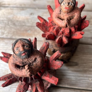 May include: Two handmade sculptures of figures with hands clasped in prayer. The figures have dark hair and are atop a spiky, red base. The sculptures are made of a textured, earthy material, with visible brushstrokes.