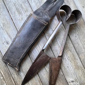 Puede incluir: Tijeras antiguas de metal con mangos en forma de corazón y una funda de cuero. Las tijeras tienen un aspecto oscuro y envejecido, con desgaste visible. La funda de cuero es de color marrón oscuro y muestra signos de edad. Las tijeras descansan sobre una superficie de madera desgastada.