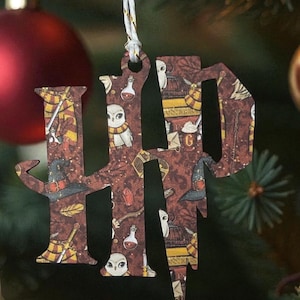 May include: Wooden Christmas ornament in the shape of the letters "HP", featuring a patterned design with owls, potions, and other magical elements. The ornament hangs from a white and gold string against a blurred Christmas tree background.