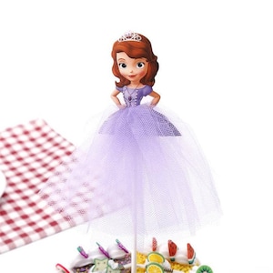 May include: Purple and white princess cake topper with a cartoon image of a princess wearing a crown and a purple dress. The princess is holding her hands on her hips. The cake topper is made of cardboard and has a tulle skirt.