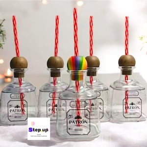 Könnte beinhalten: Fünf durchsichtige Glasflaschen mit rot-weiß gestreiften Strohhalmen, jede mit einem Korkverschluss aus Holz. Die Flaschen sind mit einer klaren Flüssigkeit gefüllt und haben ein schwarzes Etikett mit dem aufgedruckten Wort "PATRÓN". Eine Flasche hat einen regenbogenfarbenen Flaschenverschluss.