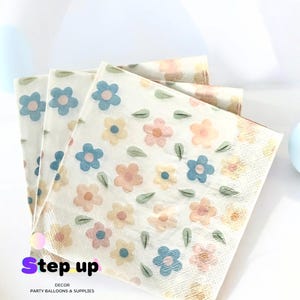 May include: A stack of white paper napkins with a floral pattern featuring blue, pink, and yellow flowers and green leaves. The napkins have the text "Step up Decor Party Balloons & Supplies" printed on the bottom.