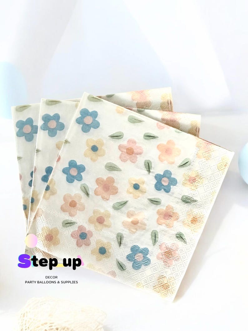 May include: A stack of white paper napkins with a floral pattern featuring blue, pink, and yellow flowers and green leaves. The napkins have the text "Step up Decor Party Balloons & Supplies" printed on the bottom.