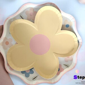 May include: A yellow and pink flower-shaped paper plate with a scalloped edge. The plate has a floral pattern with pink, yellow, and blue flowers.