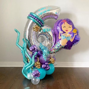 May include: A silver number six balloon with the words "Happy Birthday" printed on it. The balloon is decorated with purple, teal, and gold balloons, including a mermaid balloon with a purple tail and a gold crown.