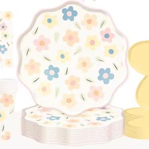 May include: A set of floral-themed party supplies, including plates, cups, and napkins. The plates feature a white background with pastel pink, yellow, and blue flowers. The cups and napkins have a similar design. The plates are stacked in a pile, and the cups and napkins are arranged in a row.