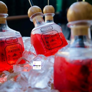 May include: Four clear glass bottles filled with a red liquid and topped with wooden stoppers. The bottles are labelled "Tequila 100% de Agave" and "Tequila Patron". The bottles are sitting in a bed of ice.