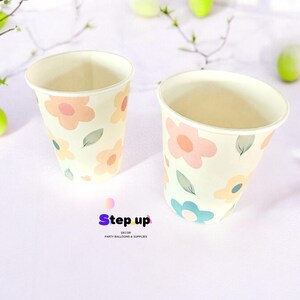 May include: Two paper cups with a floral pattern. The cups are white with pink, yellow, and blue flowers and green leaves. The cups are on a white surface. The text "Step Up Decor Party Balloons & Supplies" is at the bottom of the image.