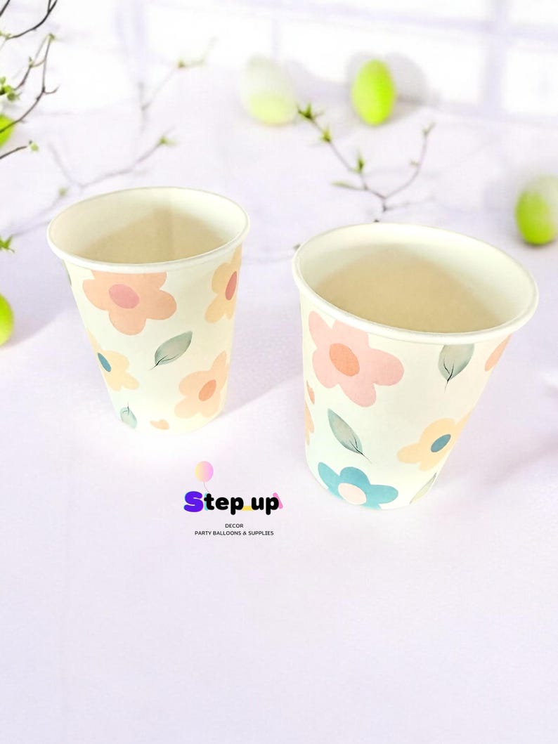 May include: Two paper cups with a floral pattern. The cups are white with pink, yellow, and blue flowers and green leaves. The cups are on a white surface. The text "Step Up Decor Party Balloons & Supplies" is at the bottom of the image.