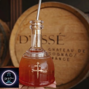 May include: A clear glass bottle with a red liquid inside. The bottle has a gold cross design on the front and a straw sticking out of the top. The bottle is in front of a wooden barrel with the text "D'SSÉ CHATEAU DE COGNAC/FRANCE" on it.