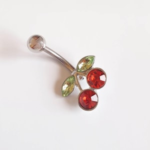 May include: A silver-coloured belly button ring with a cherry design. The cherries are red, faceted gemstones, and the leaves are green. The curved bar has a small, round ball at the top.