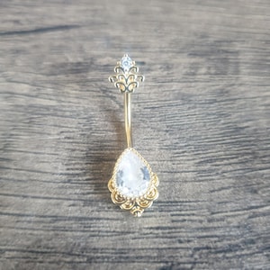 May include: A gold-coloured belly button ring featuring a teardrop-shaped, clear gemstone. The ring has an ornate design around the gemstone and a small, clear gemstone at the top. The jewellery is set against a wooden background.