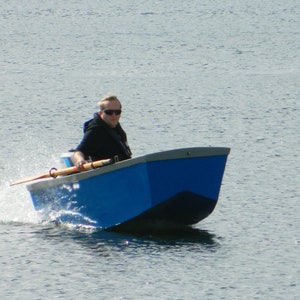 Puede incluir: Una persona rema en un bote de remos azul y gris en un lago. El bote avanza y crea una pequeña estela.