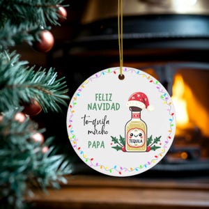 May include: A circular Christmas ornament with the text "Feliz Navidad Te-quila mucho Papa". The ornament features a tequila bottle wearing a Santa hat, surrounded by colourful lights and holly.