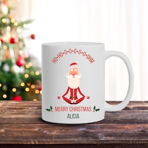 May include: White ceramic Christmas mug featuring a cartoon Santa Claus in a yoga pose. The mug has the text "HO-HO-O-O-O-OHM" and "MERRY CHRISTMAS ALICIA" printed on it. The mug is on a wooden surface with a Christmas tree in the background.