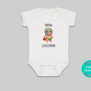May include: White baby onesie with a green, red, and blue cartoon wrestler wearing a mask. The text "little luchador" is printed on the onesie.
