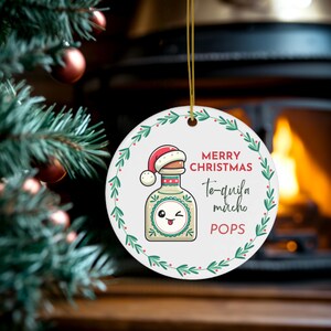 May include: A white ceramic Christmas ornament featuring a tequila bottle wearing a Santa hat. The ornament has the words "MERRY CHRISTMAS Te-quila mucho POPS" in black script and is trimmed with green leaves and red berries.