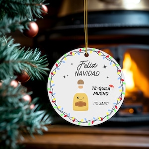 May include: Circular white Christmas decoration with a gold string, displaying the words "Feliz Navidad" and "Te-quila Mucho" alongside a cartoon tequila bottle and a Santa hat. The ornament is framed by a string of colourful lights.