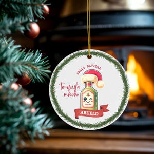 May include: A circular Christmas tree decoration featuring a tequila bottle wearing a Santa hat. The ornament has the words "Feliz Navidad", "Te-quila mucho", and "Abuelo". A green wreath border surrounds the design.
