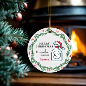 May include: A circular white Christmas ornament with a green wreath and red berry design. The ornament has the text "MERRY CHRISTMAS te-quila mucho Abuelo" and a cartoon tequila bottle wearing a Santa hat.