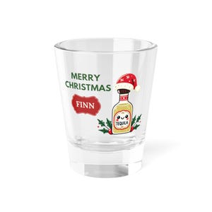 May include: A clear shot glass with the text "MERRY CHRISTMAS FINN" in green and red. The glass features a cartoon tequila bottle wearing a Santa hat and holly leaves. The word "TEQUILA" is on the bottle.