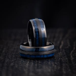 May include: Two black tungsten carbide rings with blue inlay. The rings are stacked on top of each other.