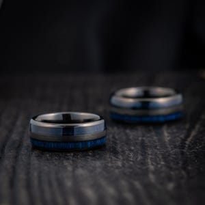 May include: Two black and blue tungsten carbide rings with a brushed finish. The rings have a blue inlay and are stacked on top of each other.