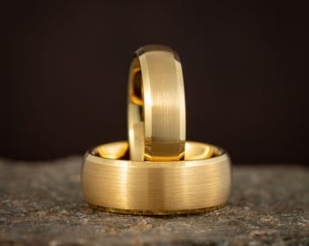 May include: Two gold wedding bands, one resting atop the other, are displayed on a textured stone surface. The rings have a brushed finish and a rounded profile. The background is a dark, solid color.