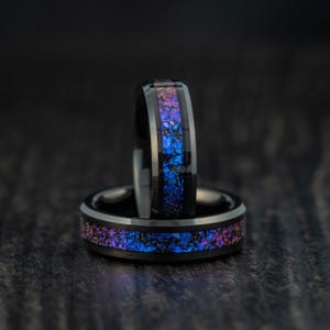 May include: Two black rings with a vibrant, colorful inlay of blue, purple, and pink. The rings are set against a dark background, highlighting the iridescent design. The rings are likely made of a durable material.