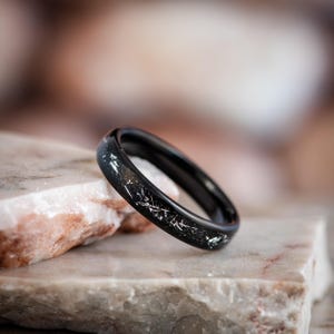 May include: A black ring with a band featuring a speckled design and silver-colored accents. The ring is set on a light-colored stone surface, with a blurred background. The ring is a piece of jewelry.