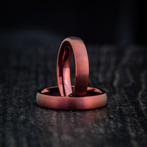 May include: A pair of rings with a brushed copper finish. One ring is positioned vertically, while the other lies flat horizontally beneath it. Both rings feature a smooth, metallic surface with a distinct reddish-brown hue and a subtle texture.