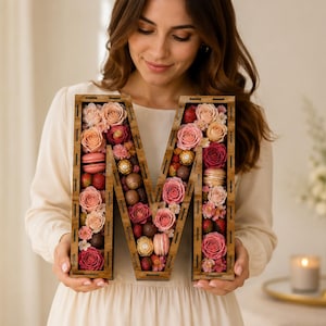 May include: A wooden letter 'M' filled with pink and red roses, macarons, strawberries, and chocolates. The letter is held by a person wearing a cream dress. A candle and vase of flowers are in the background.
