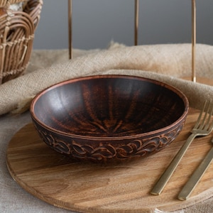 Könnte beinhalten: Eine rustikale Tischdekoration mit einer braunen Keramikschale mit dekorativem Rand, einem Holzservierbrett und goldenem Besteck. Drei weiße Kerzen in goldenen Haltern stehen hinter der Schale. Ein Weidenkorb mit Brot befindet sich im Hintergrund.