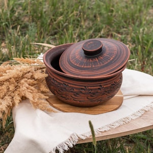Può includere: Una casseruola in ceramica marrone con coperchio, appoggiata su un sottopentola in legno e un panno di lino con frange. Il piatto presenta un motivo floreale decorativo. La scena è ambientata all'aperto con erba alta sullo sfondo.