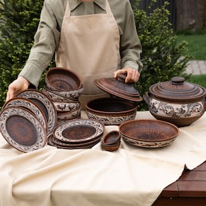 Op de afbeelding: Een verzameling handgemaakt, rustiek bruin keramisch servies, waaronder borden, kommen en een afgedekte ovenschaal. De stukken hebben een decoratief wit bloemenpatroon rond de randen. De set is gerangschikt op een beige tafelkleed.
