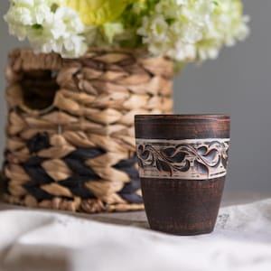 Puede incluir: Una taza de cerámica marrón oscuro con una banda blanca con un diseño floral tallado. Una cesta tejida con un patrón negro y marrón y un ramo de flores blancas y un anturio amarillo en el fondo.