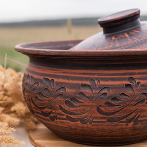 Puede incluir: Una olla de cerámica marrón con tapa, que presenta un diseño floral decorativo. La olla está sobre una superficie de madera y un paño blanco.