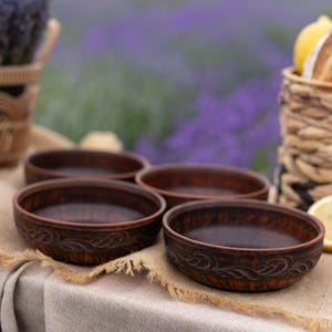 May include: Four brown ceramic bowls with decorative floral patterns sit on a beige linen tablecloth. The bowls are round with a slightly raised rim. The background features a lavender field and a basket with lemons.