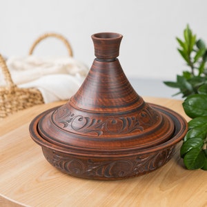Puede incluir: Una tagine de cer&aacute;mica marr&oacute;n con intrincados dise&ntilde;os florales. La tagine est&aacute; sobre una mesa de madera.