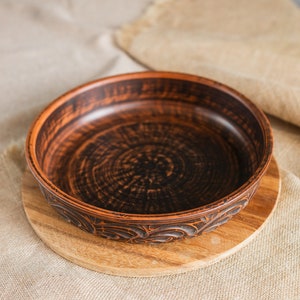 Puede incluir: Un cuenco de cerámica marrón hecho a mano con un borde decorativo, apoyado sobre una tabla de cortar de madera. El cuenco tiene un interior oscuro y una superficie texturizada. El cuenco tiene unos 25 cm de diámetro, perfecto para servir comida.